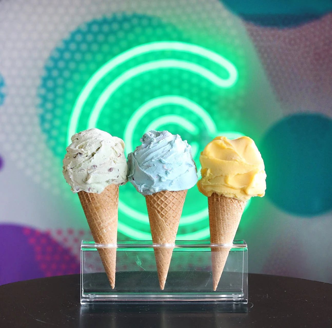 Three scoops of ice cream in waffle cones placed on a clear stand against a colorful, blurred background with a green Wi-Fi symbol.