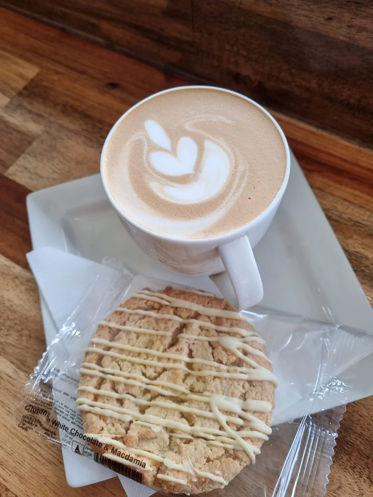 A cup of coffee with latte art on top and a wrapped oatmeal cookie with white chocolate drizzle on a white square plate.