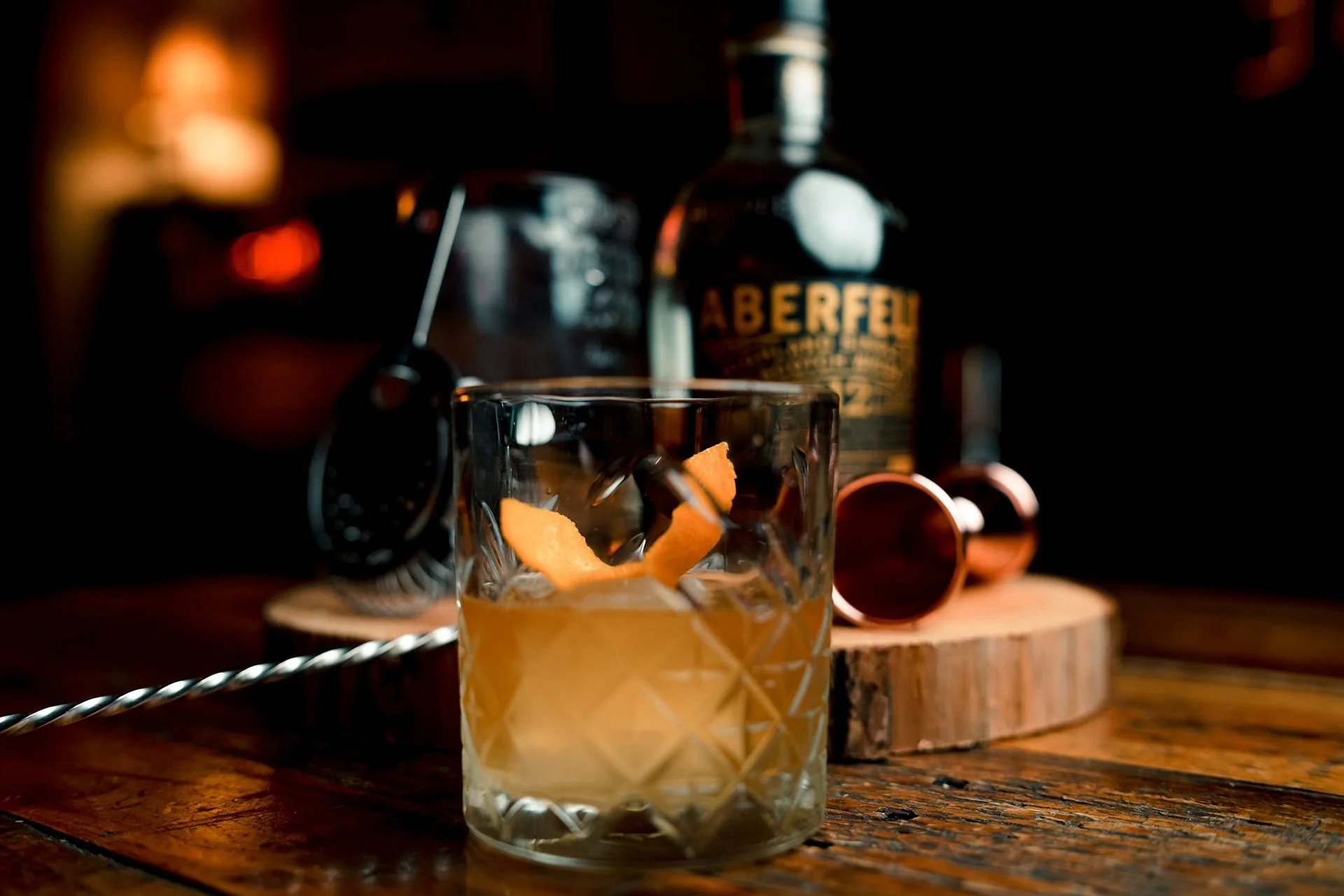A glass of whiskey with ice and orange peel garnish on a wooden table, with a bottle of Aberfeldy whiskey and a rotary phone in the background.