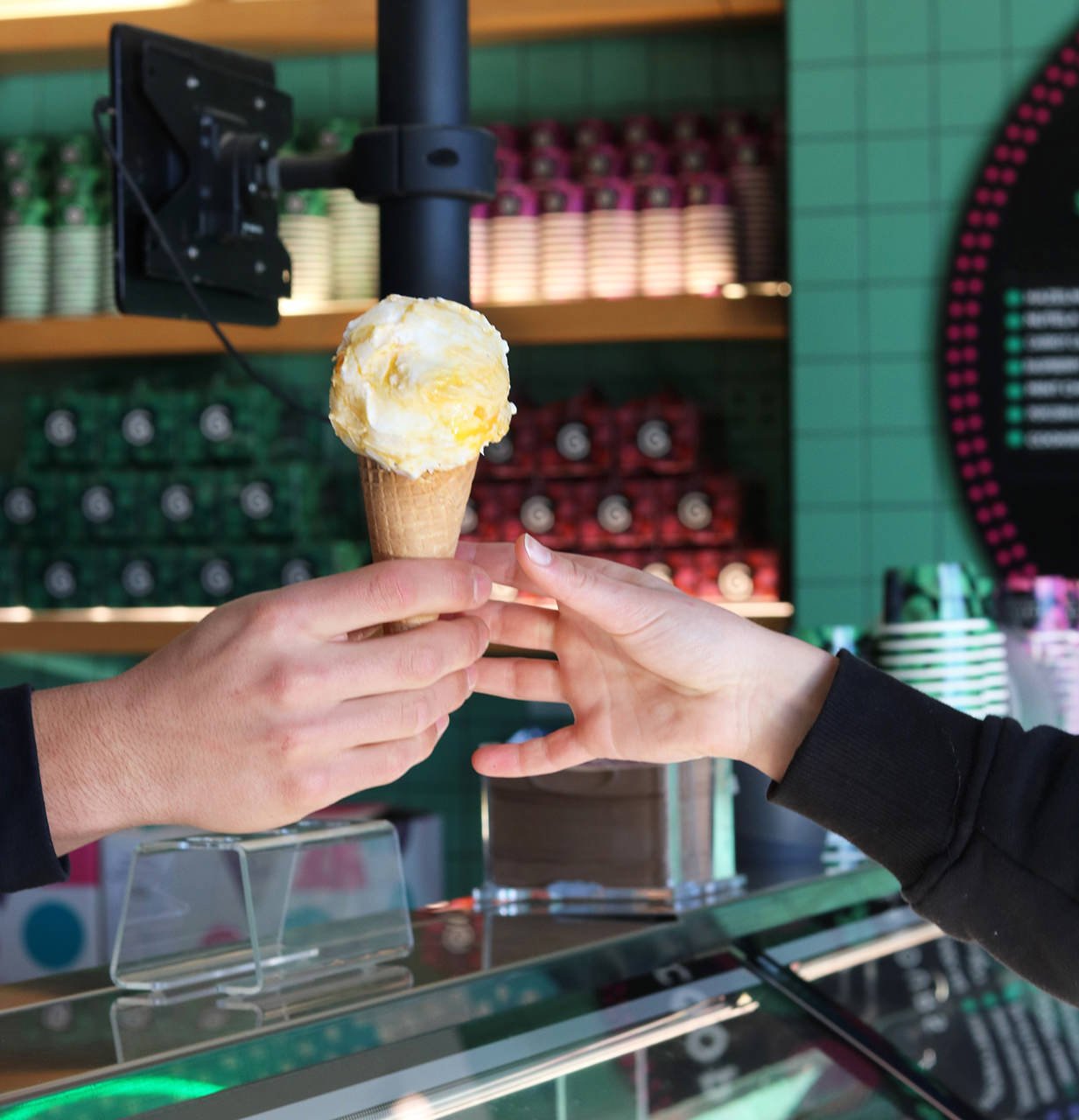 Two hands exchanging a scoop of vanilla ice cream in a waffle cone in front of a colorful store display.