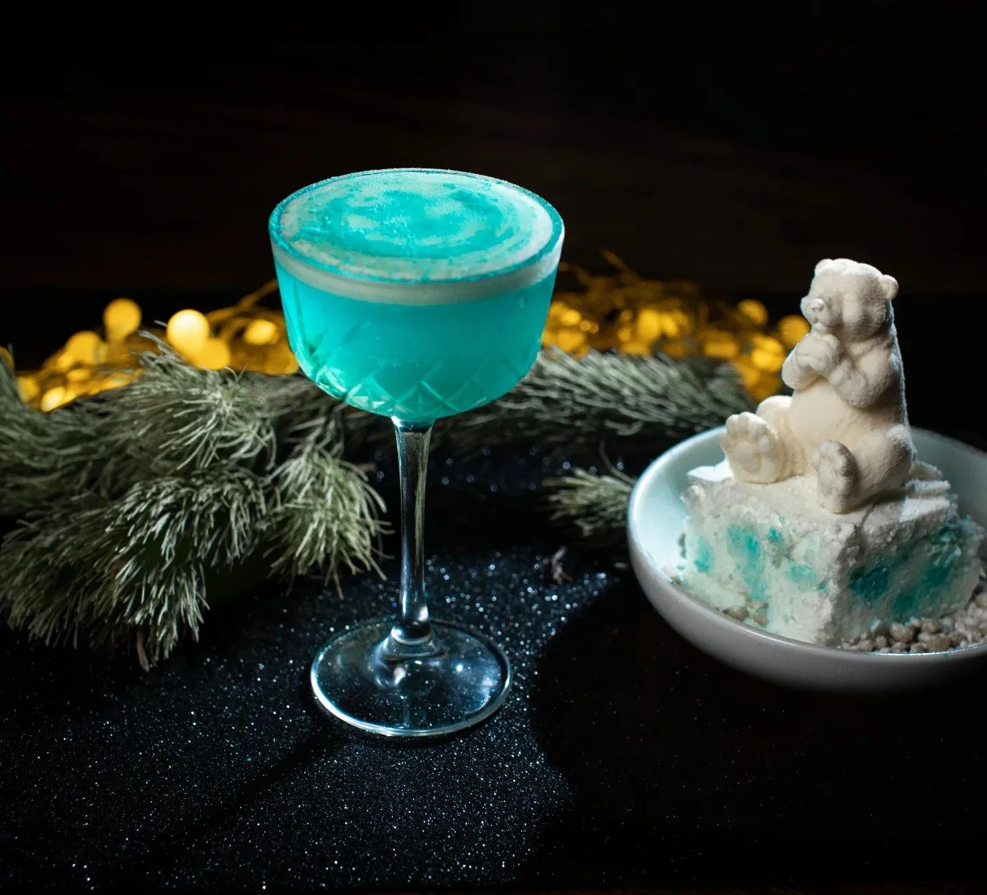 A blue cocktail in a martini glass, a small white teddy bear figurine on a dessert or ice cream in a white bowl, with pine branches and golden lights in the background.
