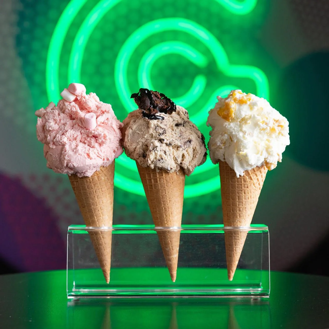 Three scoops of ice cream in cones, with pink, chocolate with toppings, and vanilla flavors, on a transparent stand against a background with a green neon sign.
