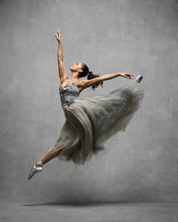 Ballet dancer in mid-air with arms extended, wearing a tutu and point shoes against a plain gray background.