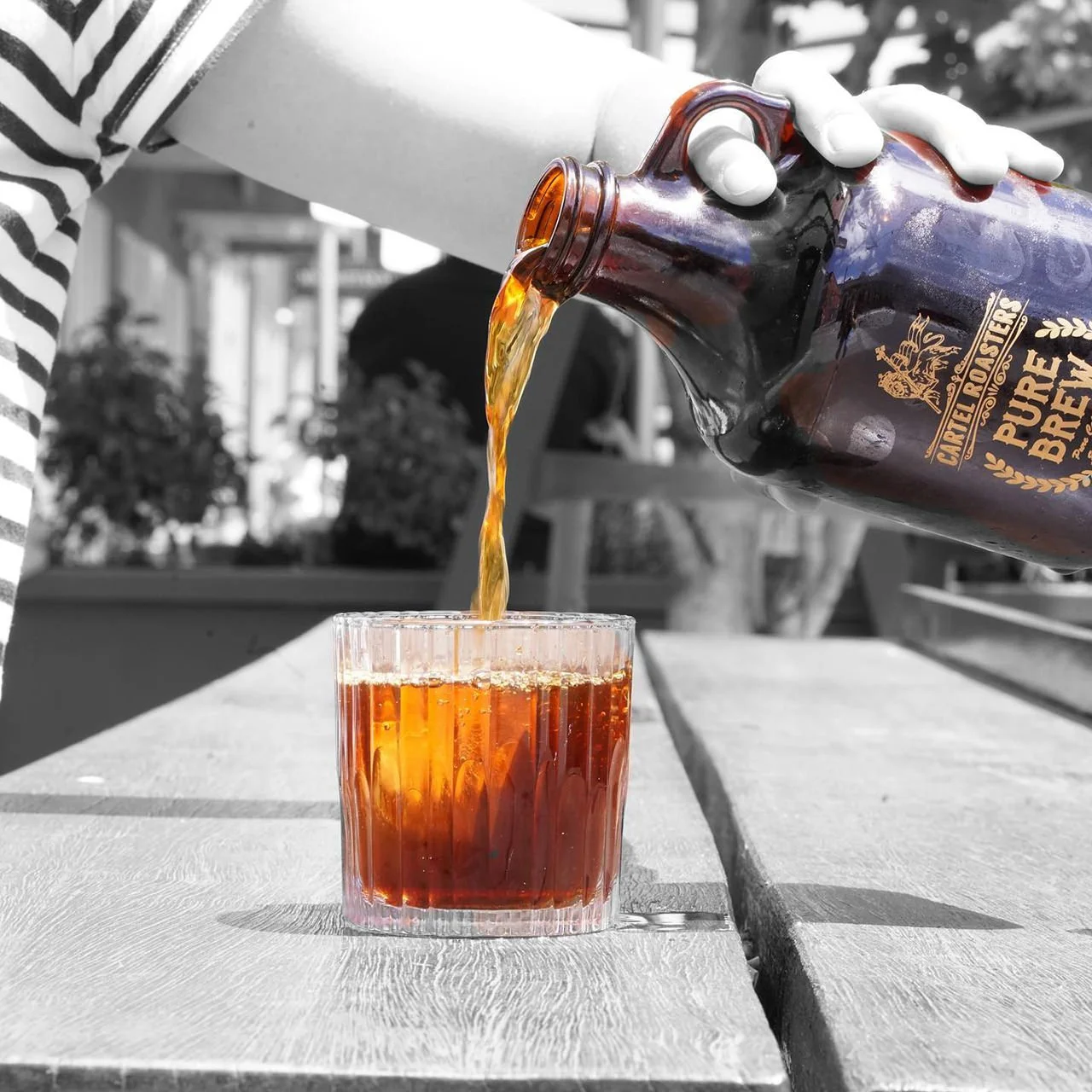 A person pouring a dark beer from a brown bottle labeled 'Carl Roaster' into a glass on a wooden table. The image is in black and white, except for the beer and bottle which are in color.