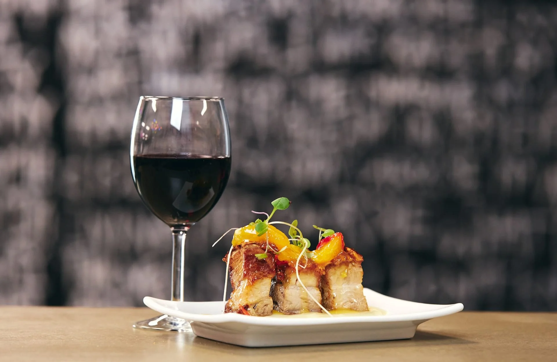 A glass of red wine next to a plated serving of cooked pork belly topped with yellow and red peppers and microgreens, on a wooden table against a gray textured background.