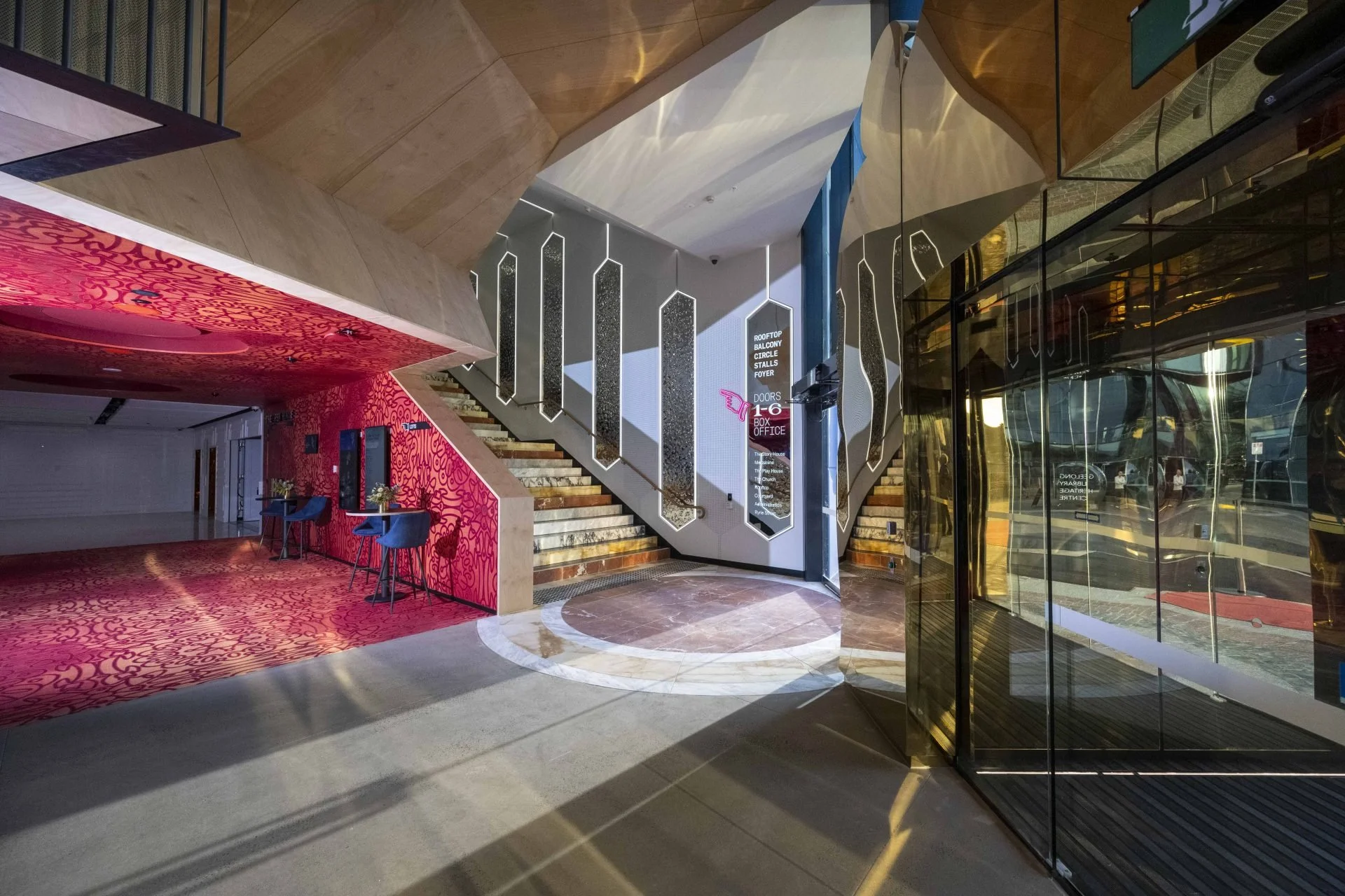Interior view of a modern building lobby with a staircase, mirrored walls, a digital directory, and seating area with blue chairs, illuminated by natural sunlight.