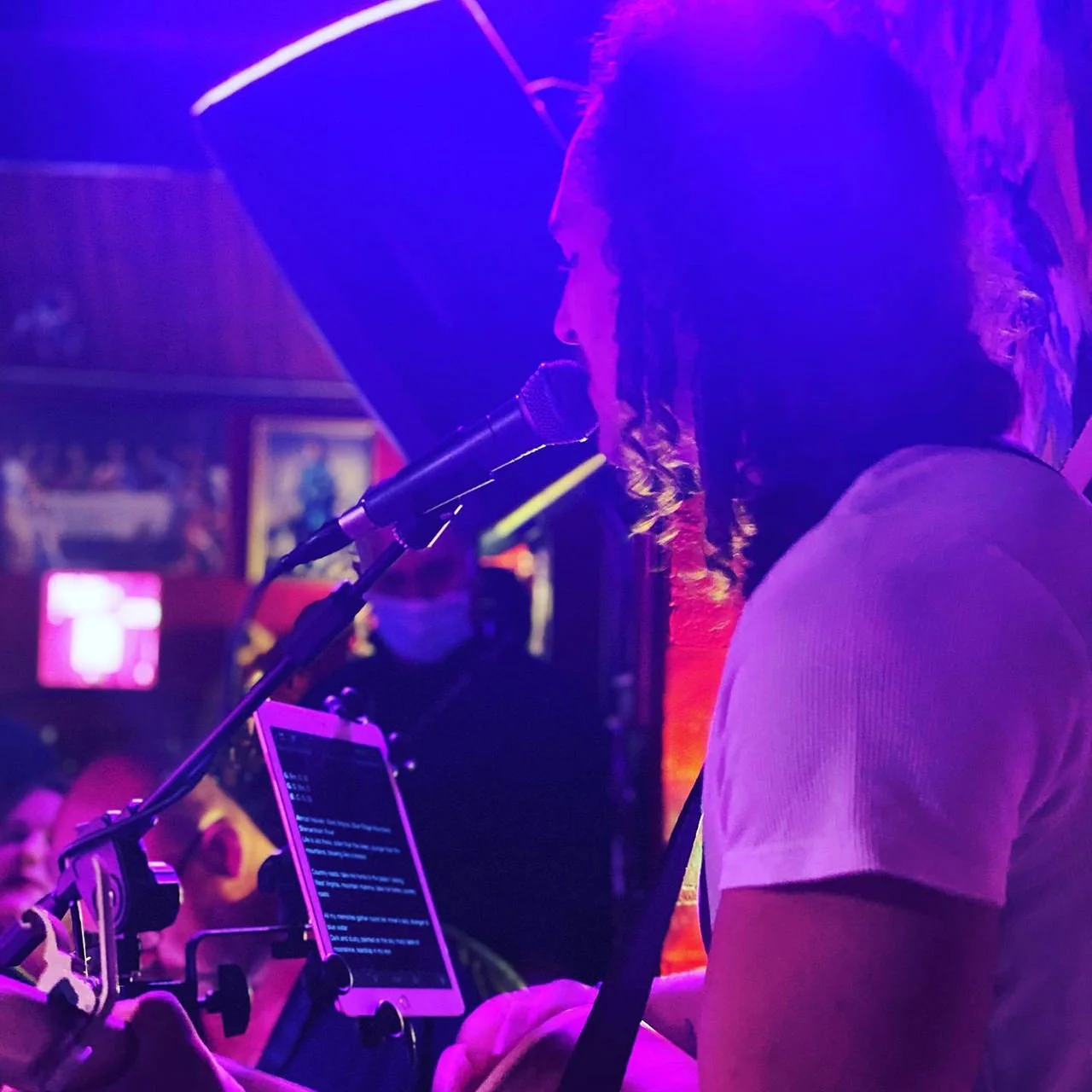 A person with dreadlocks singing into a microphone on stage, illuminated by purple and pink lighting, with a tablet in front showing lyrics.