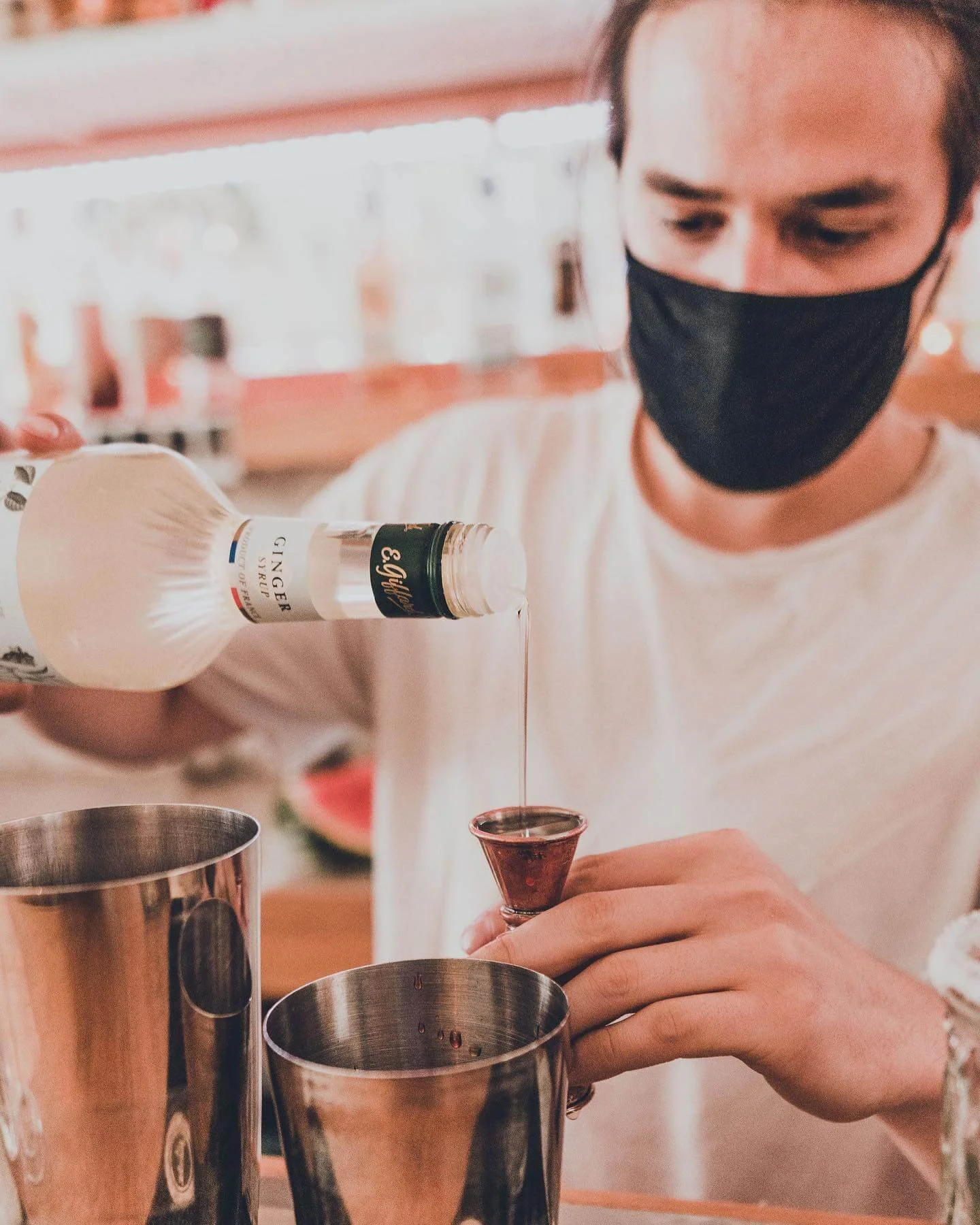 A man wearing a black mask and a white shirt pouring gin into a small glass at a bar, with metallic shakers nearby.