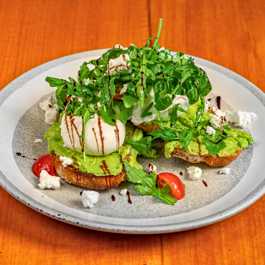 Plate of avocado toast topped with leafy greens, cherry tomatoes, poached egg, crumbled cheese, and balsamic glaze, on a speckled ceramic plate.