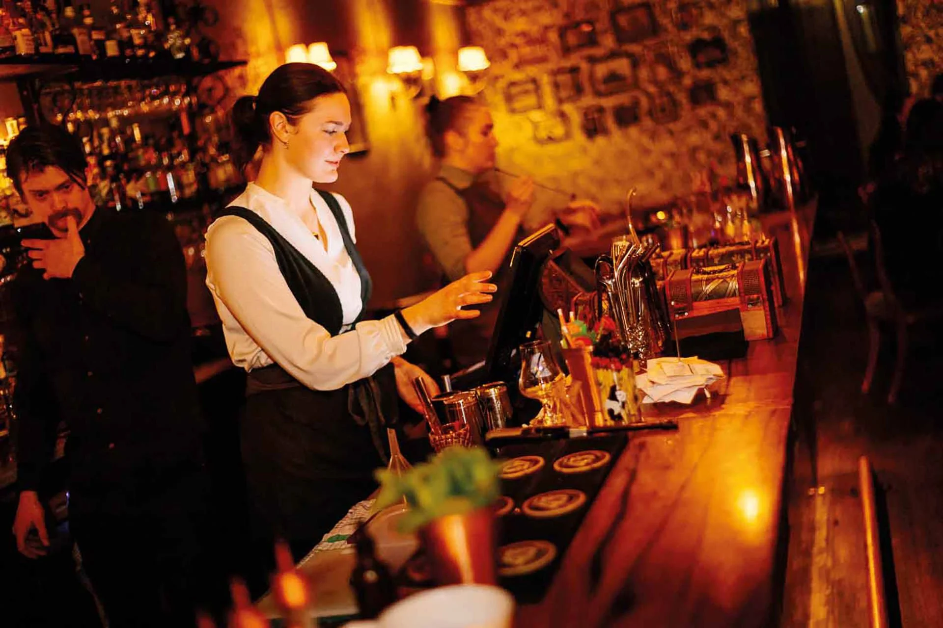 A bartender in a white shirt and black apron working behind a wooden bar counter at a dimly lit bar or pub, with a man in a black jacket and a woman in a gray top in the background.