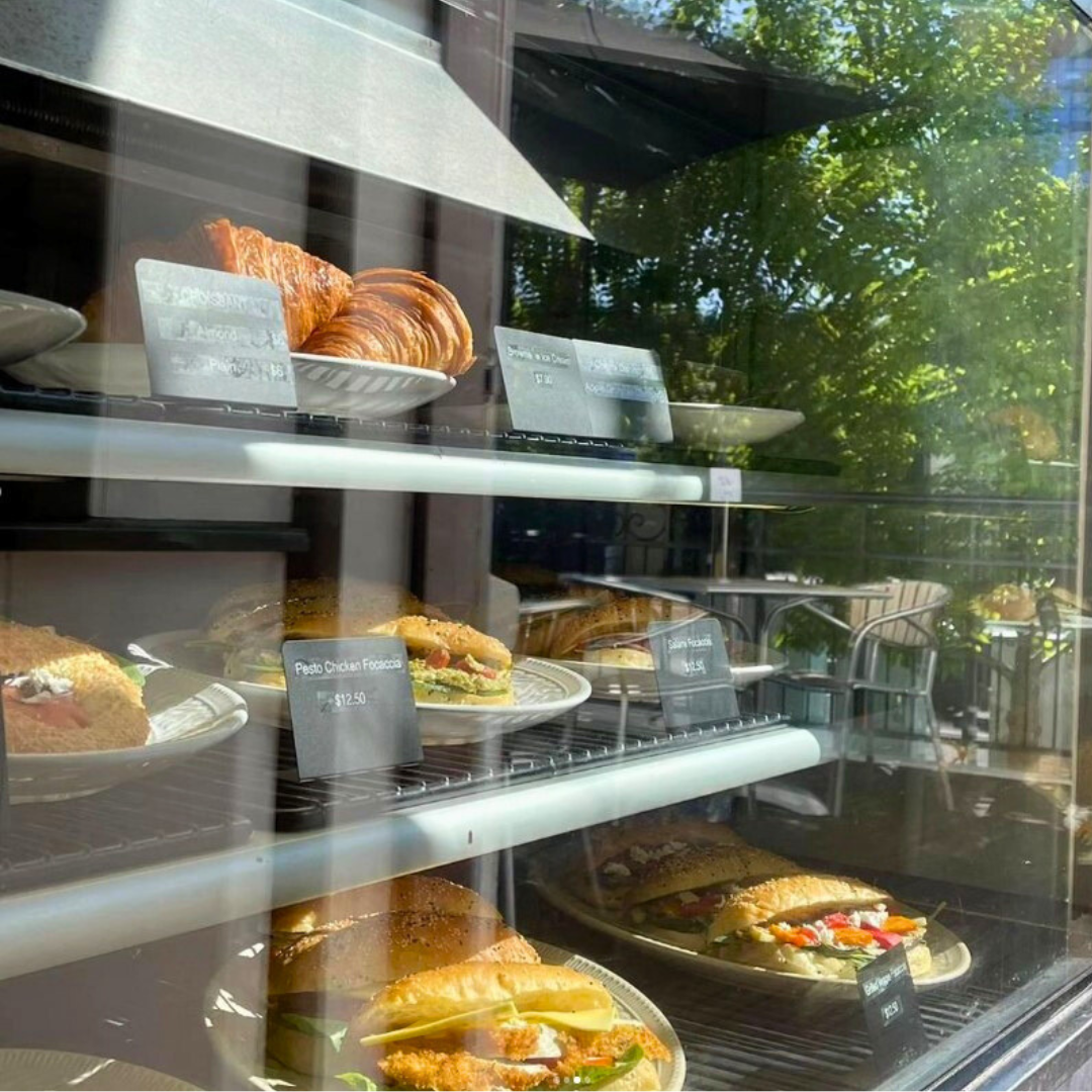 Display case with croissants, sandwiches, and pastries at a bakery or cafe, seen through a glass window with reflections of trees outside.
