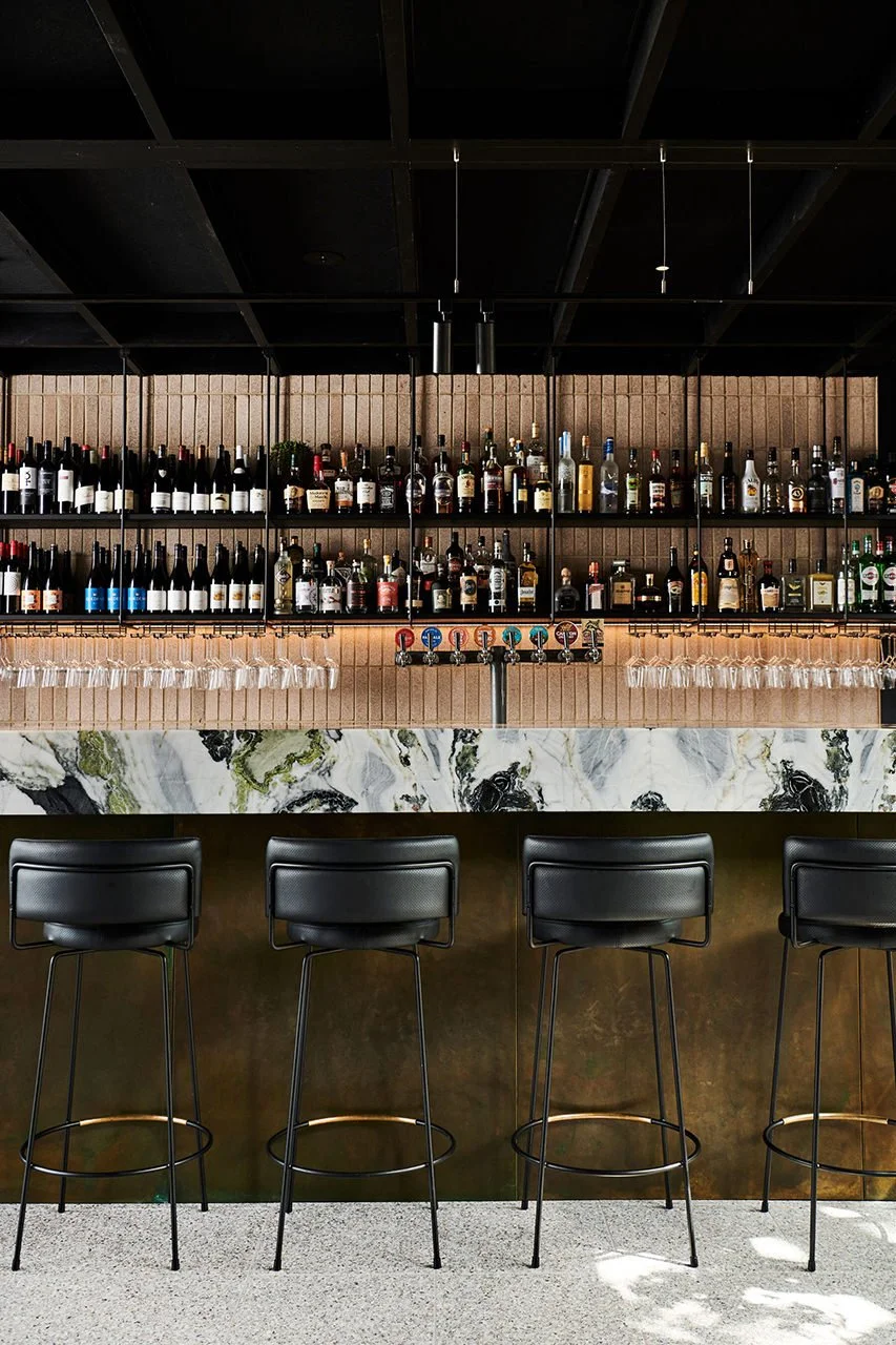 A modern bar with four black high chairs, a marble countertop, and a back wall stocked with various bottles of alcohol and hanging wine glasses.