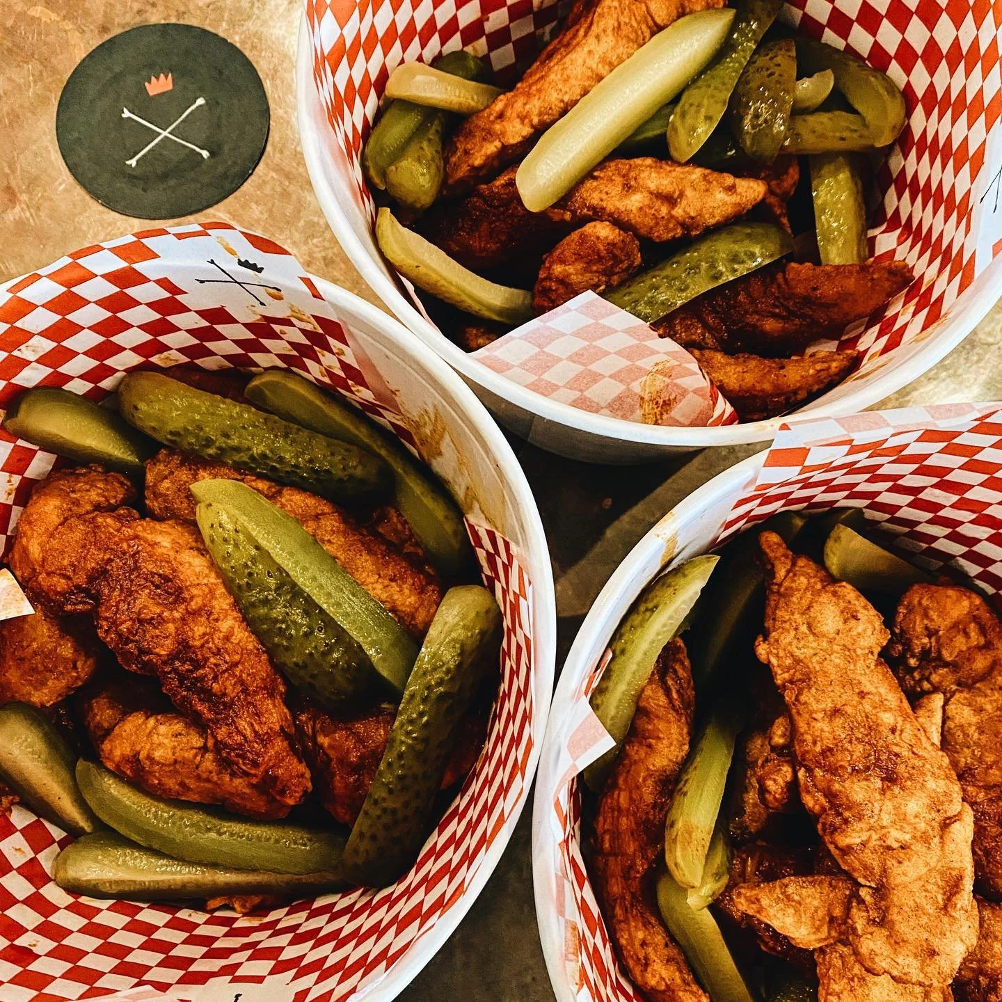Three baskets of fried chicken wings with pickles on top, lined with red and white checkered paper.