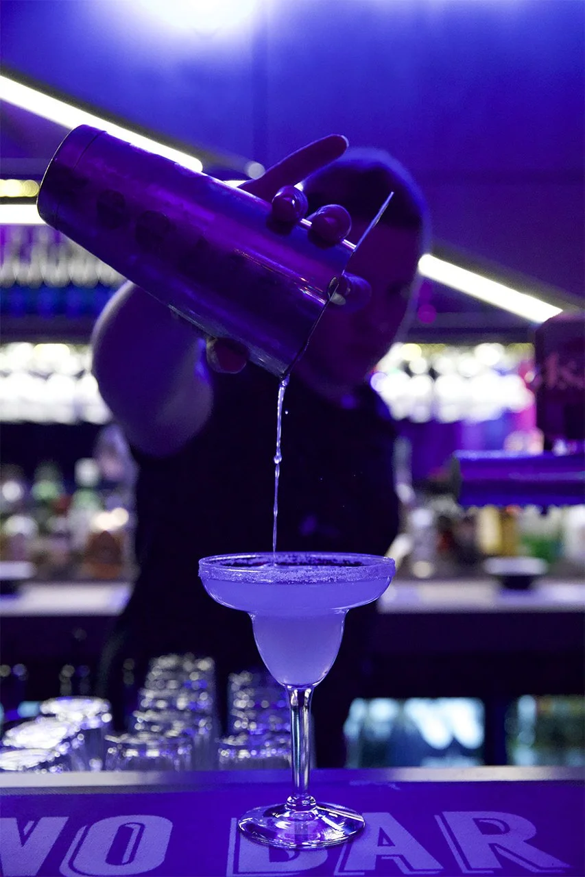 A bartender pouring a cocktail into a glass in a neon-lit bar with purple and blue lighting.