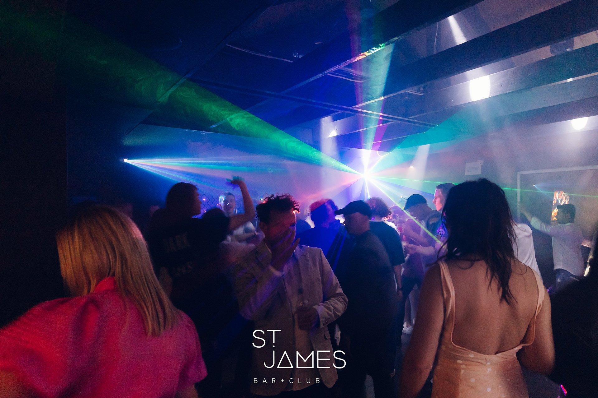 People dancing and socializing in a nightclub with colorful laser lights and fog, with a sign reading 'St. James Bar + Club' at the bottom.