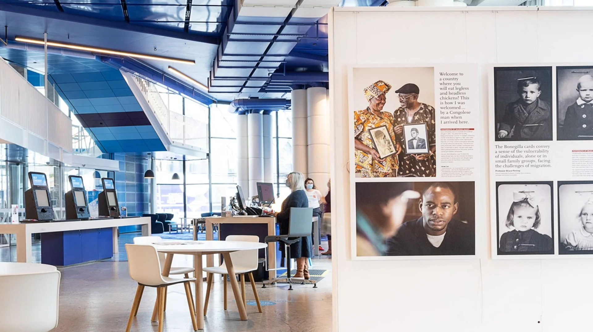 An airport or train station waiting area with check-in kiosks, a woman in a mask standing at a desk with a computer, and an exhibit on the wall displaying black and white and color photographs of different people.