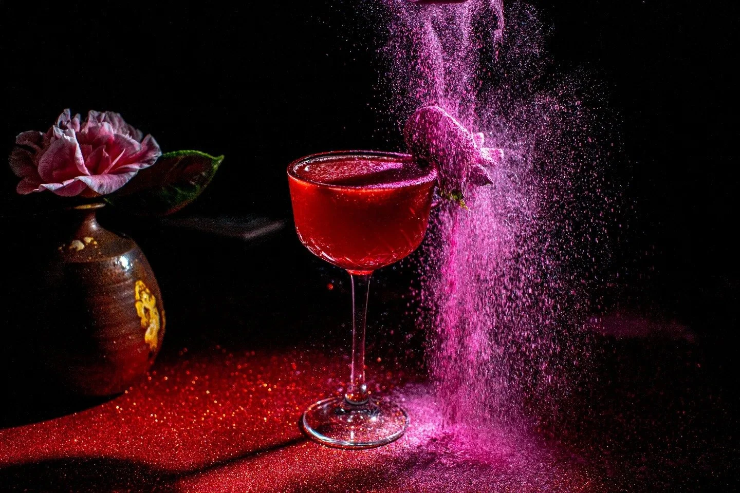 A pink cocktail in a glass with pink powder exploding into the air, with a pink flower in a brown vase on a dark background.