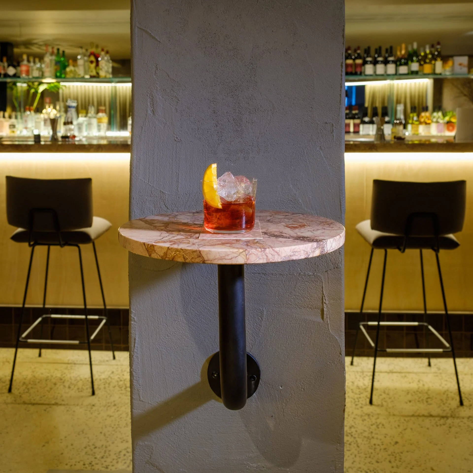 A cocktail with ice and a lemon wedge on a marble table in a bar setting.