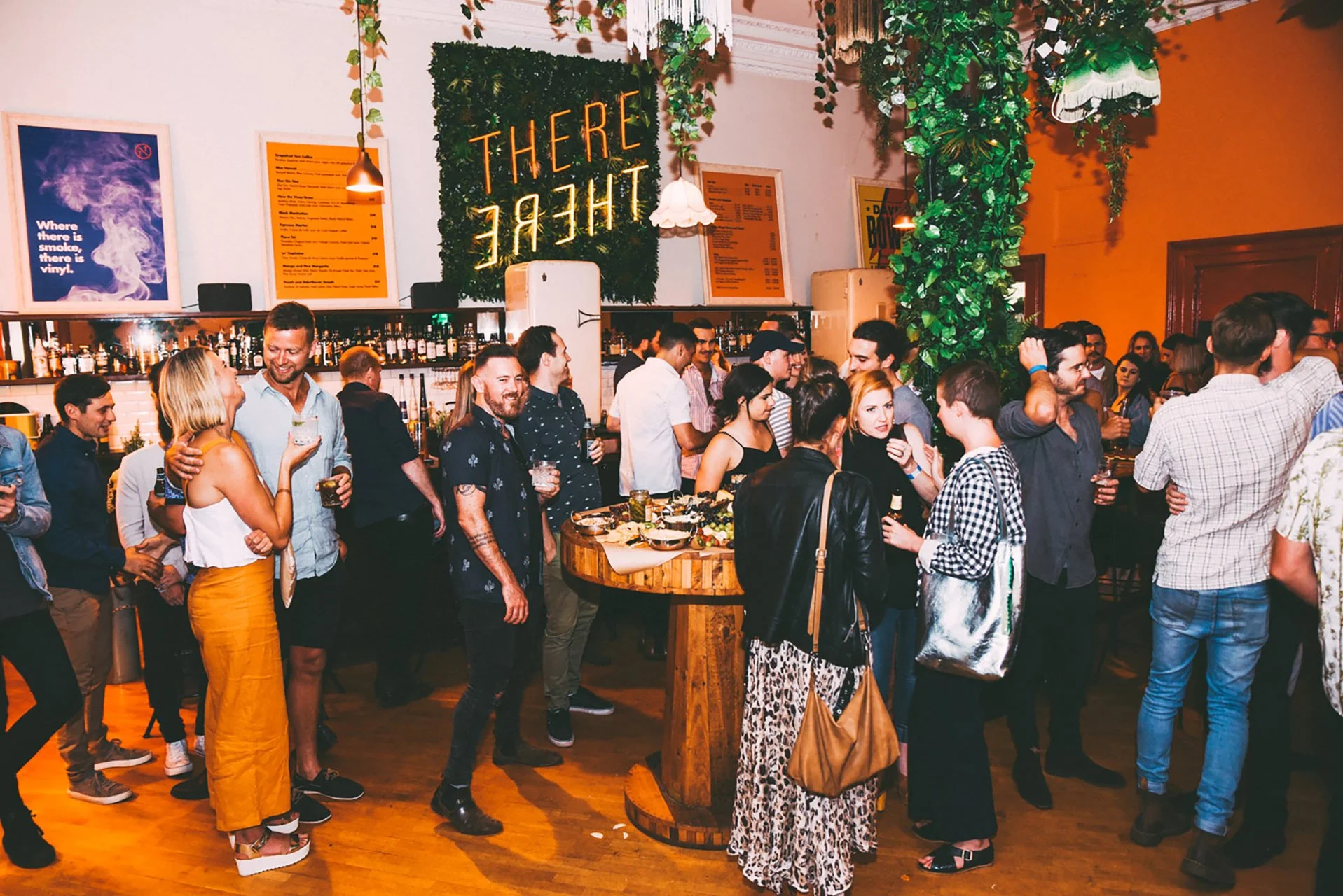 Crowd of people gathered in a bar or club with warm lighting, decorated with greenery and a neon sign that says 'THERE.'