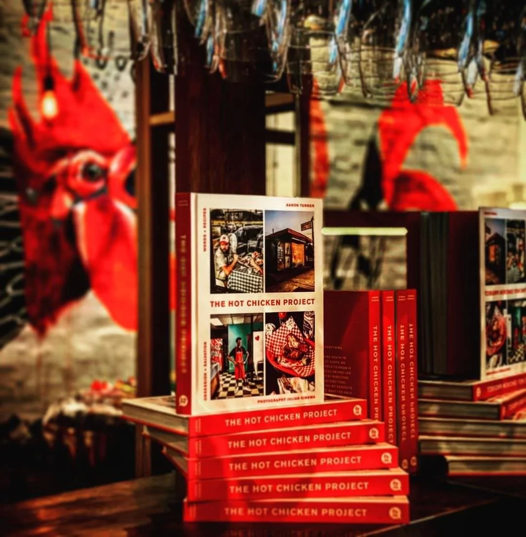 Stack of books titled 'The Hot Chicken Project' on a table in a restaurant, with hanging fish and a red chili pepper decoration in the background.