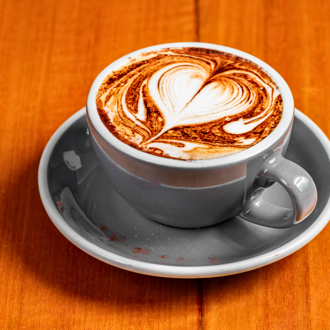 A cup of latte with chocolate and white foam art on top, served on a grey saucer on a wooden table.
