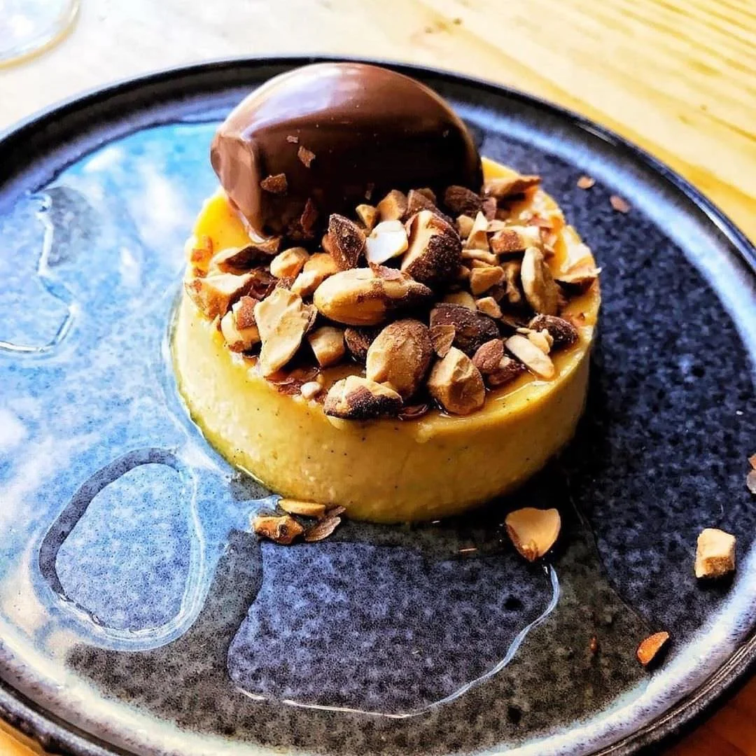 A round dessert topped with chopped nuts, a chocolate piece, and a light-colored sauce, served on a black plate.