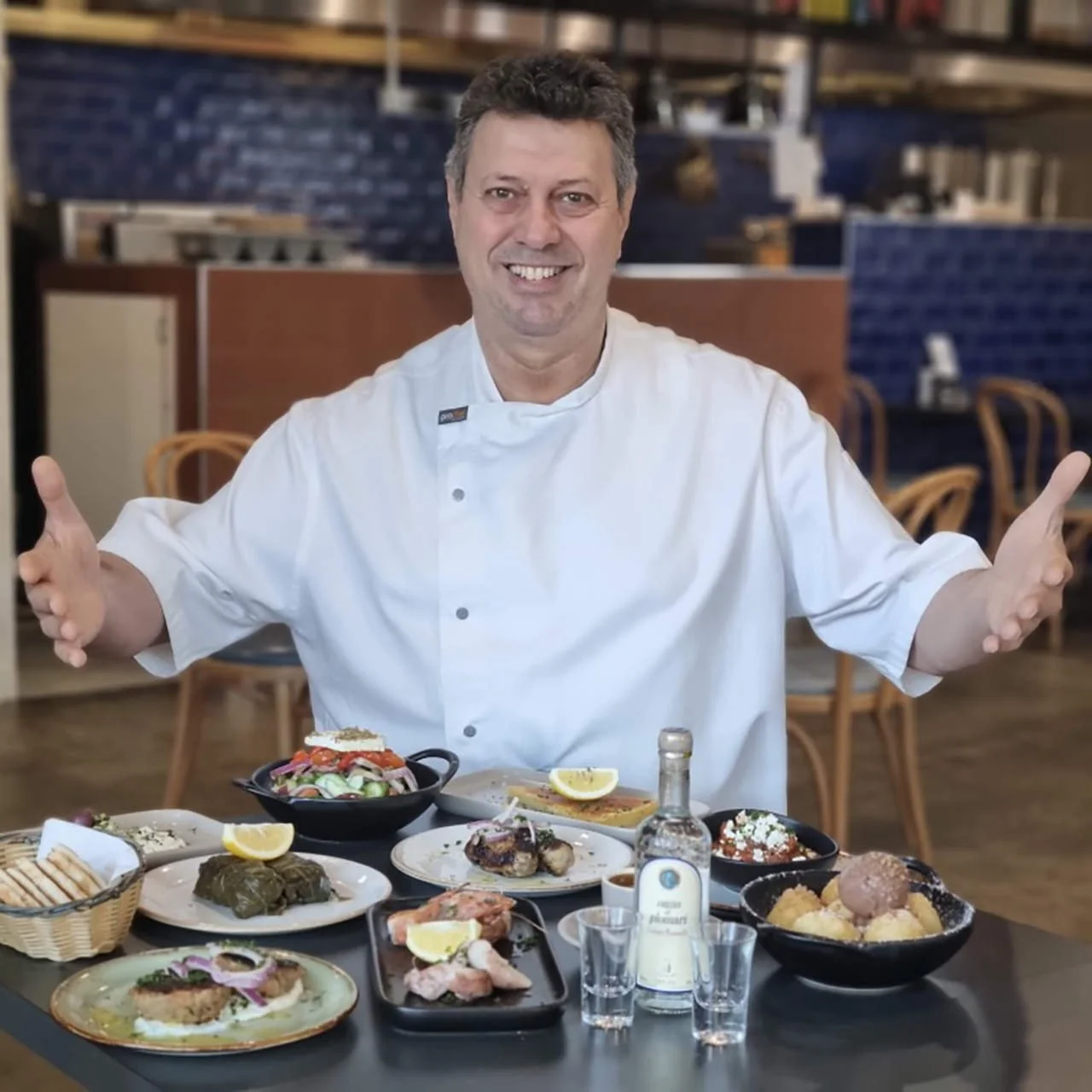 A chef in a white uniform stands behind a table filled with various dishes and bottles, smiling and gesturing with open arms in a restaurant setting.