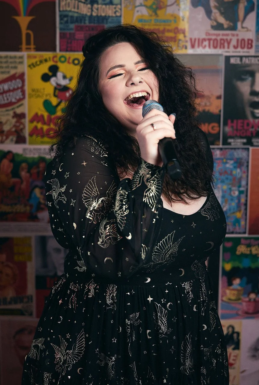 A woman with curly dark hair and light skin, singing into a microphone, smiling with her eyes closed, in front of a colorful wall decorated with retro posters.