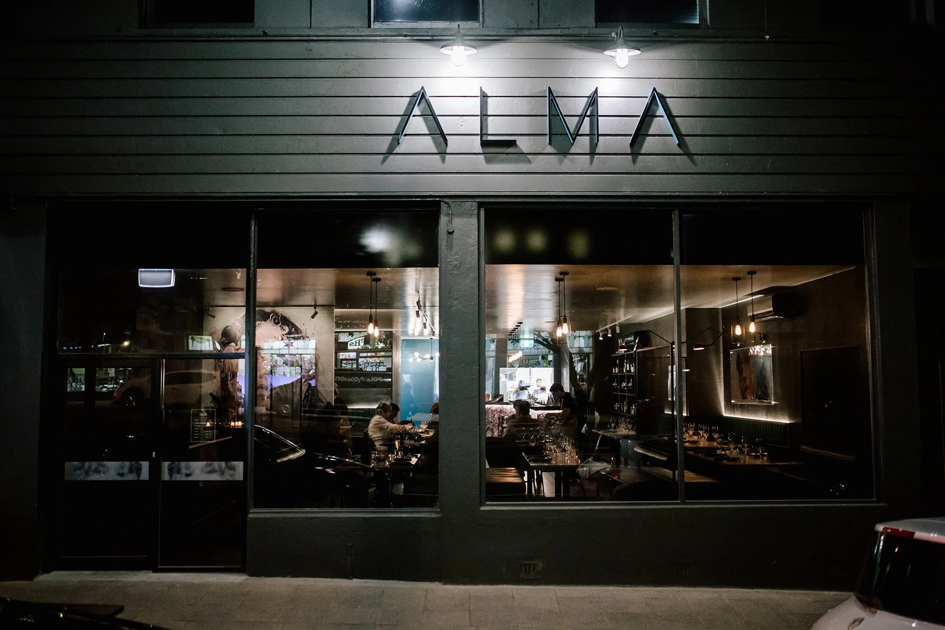 Nighttime view of a restaurant with a large window showing the interior, illuminated Sign above the entrance reads 'ALMA'.