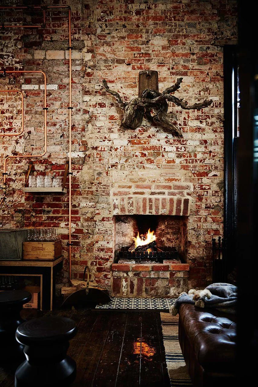 Cozy living room with a brick fireplace, a fire burning, a leather couch with a blanket, and rustic decor including a driftwood wall art piece, copper pipes, and glasses on a shelf.