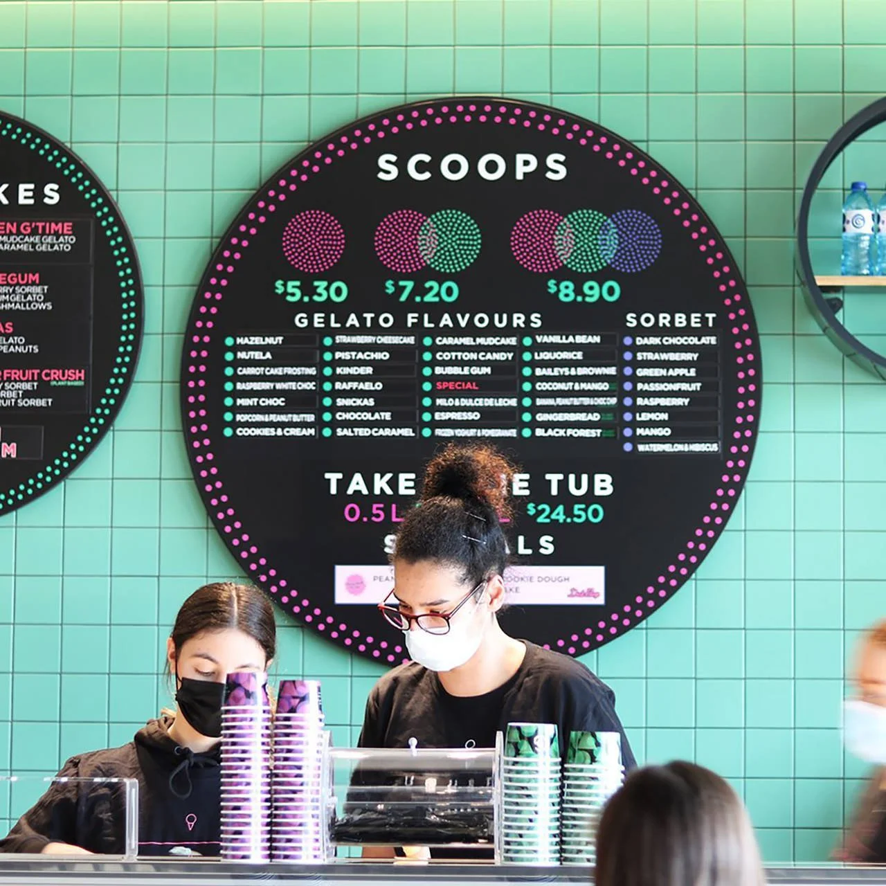 A menu board displaying gelato scoop prices, flavors, and toppings in a gelato shop, with two employees behind the counter wearing face masks.