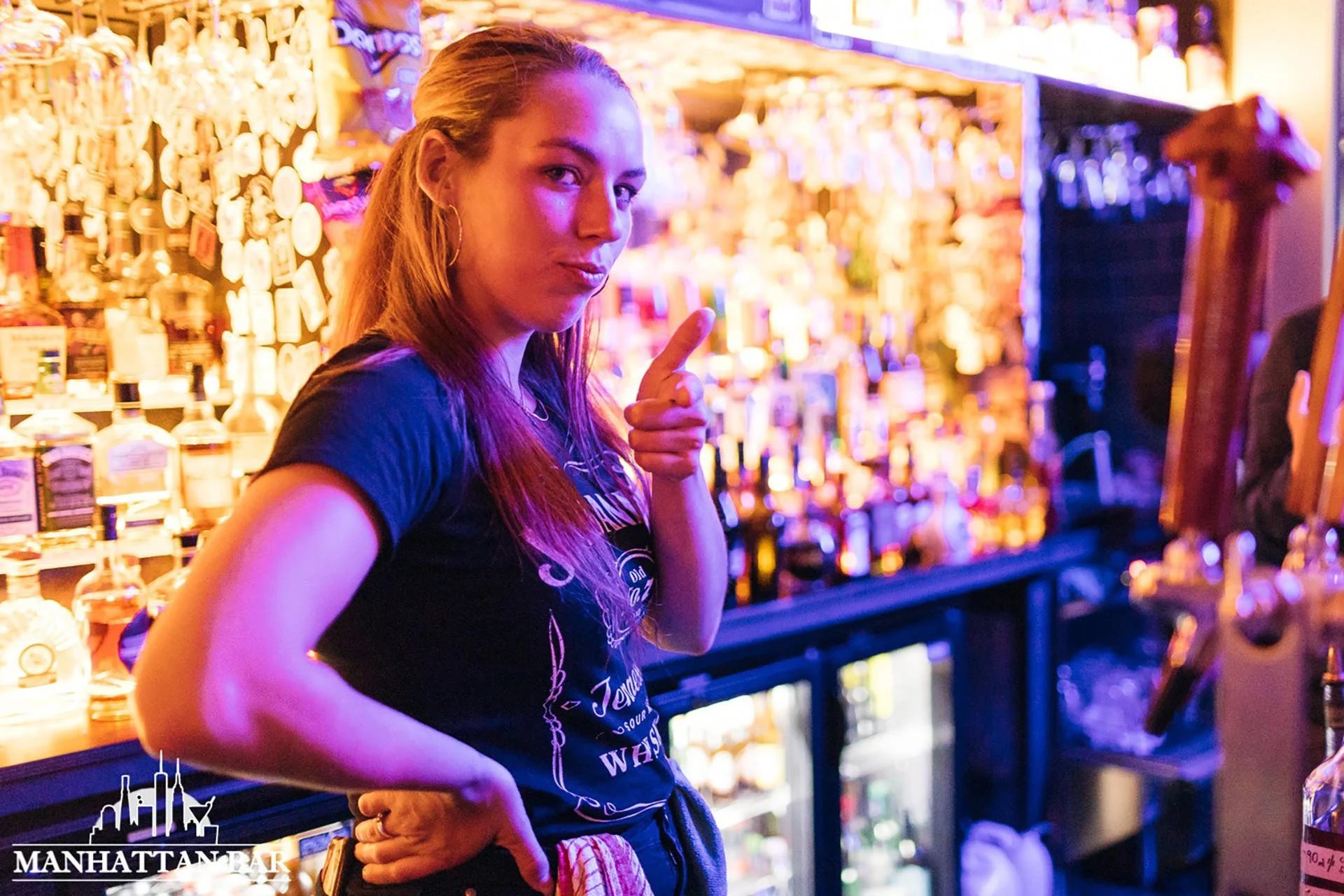 A woman with long red hair standing behind a bar, pointing with her right hand and looking at the camera, in a dimly lit bar with shelves of liquor bottles illuminated in the background.