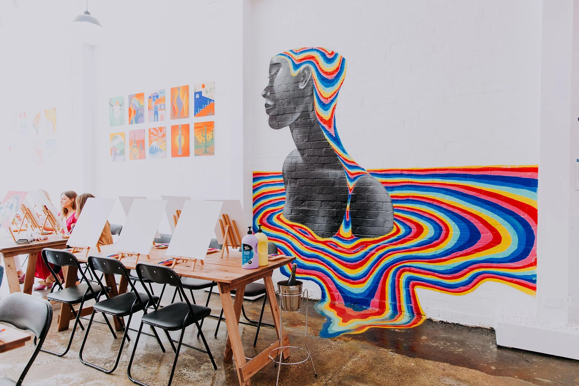 Indoor art studio with a colorful mural of a woman's profile with flowing hair on a white brick wall. The mural features vibrant, swirling colors. There are tables with blank canvases, and two women working on artwork.
