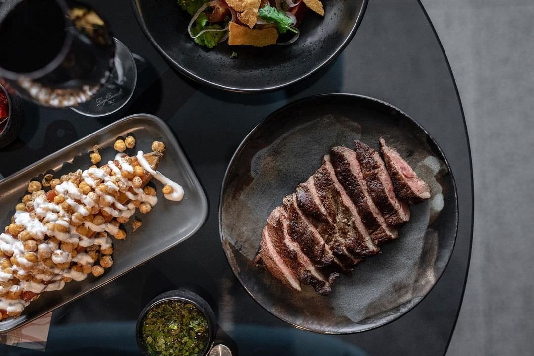 A top-down view of a black table with dishes including sliced steak on a black plate, a tray of fried food with white sauce, a salad, and two glasses of red wine.