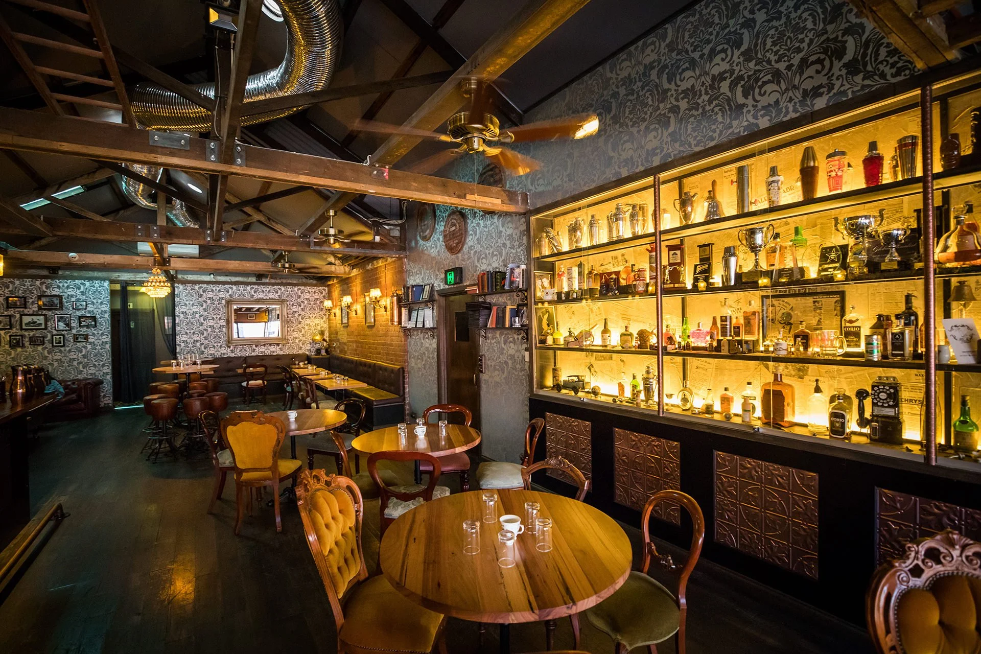 Interior of a cozy bar or restaurant with wooden furniture and vintage decor, featuring a well-lit display of bottles and trophies on shelves, patterned wallpaper, and dim warm lighting.