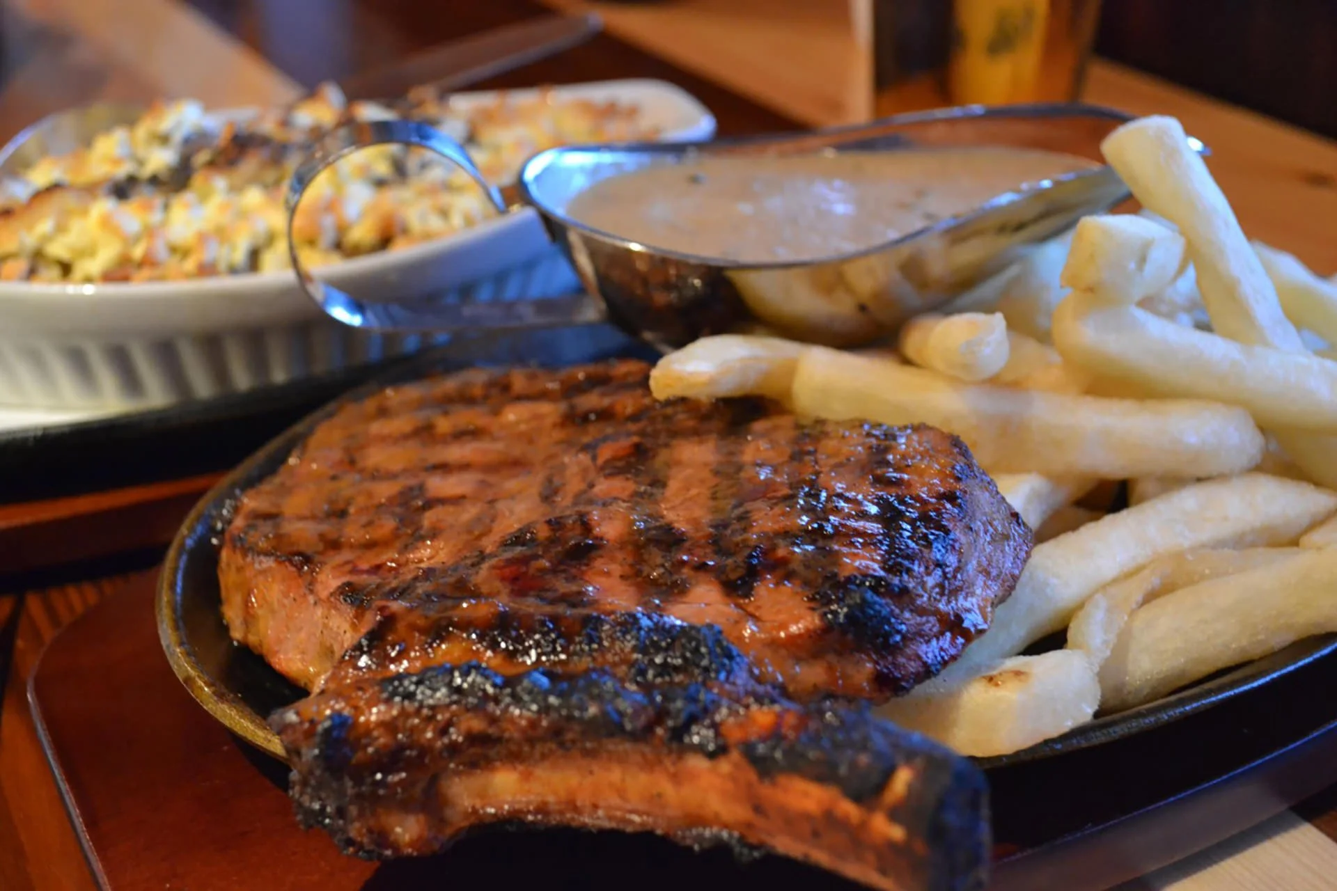 Grilled pork chop with French fries and gravy, with a side dish of rice and vegetables in the background.