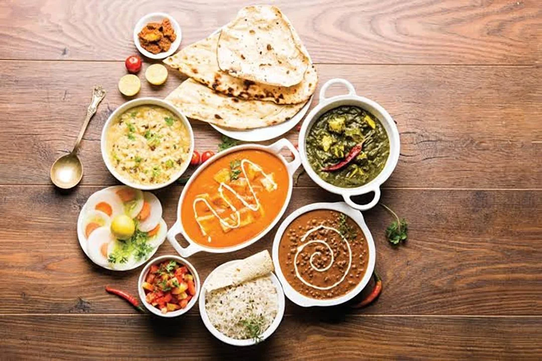 Indian meal served on a wooden table, including naan bread, various curries, rice, and side dishes.