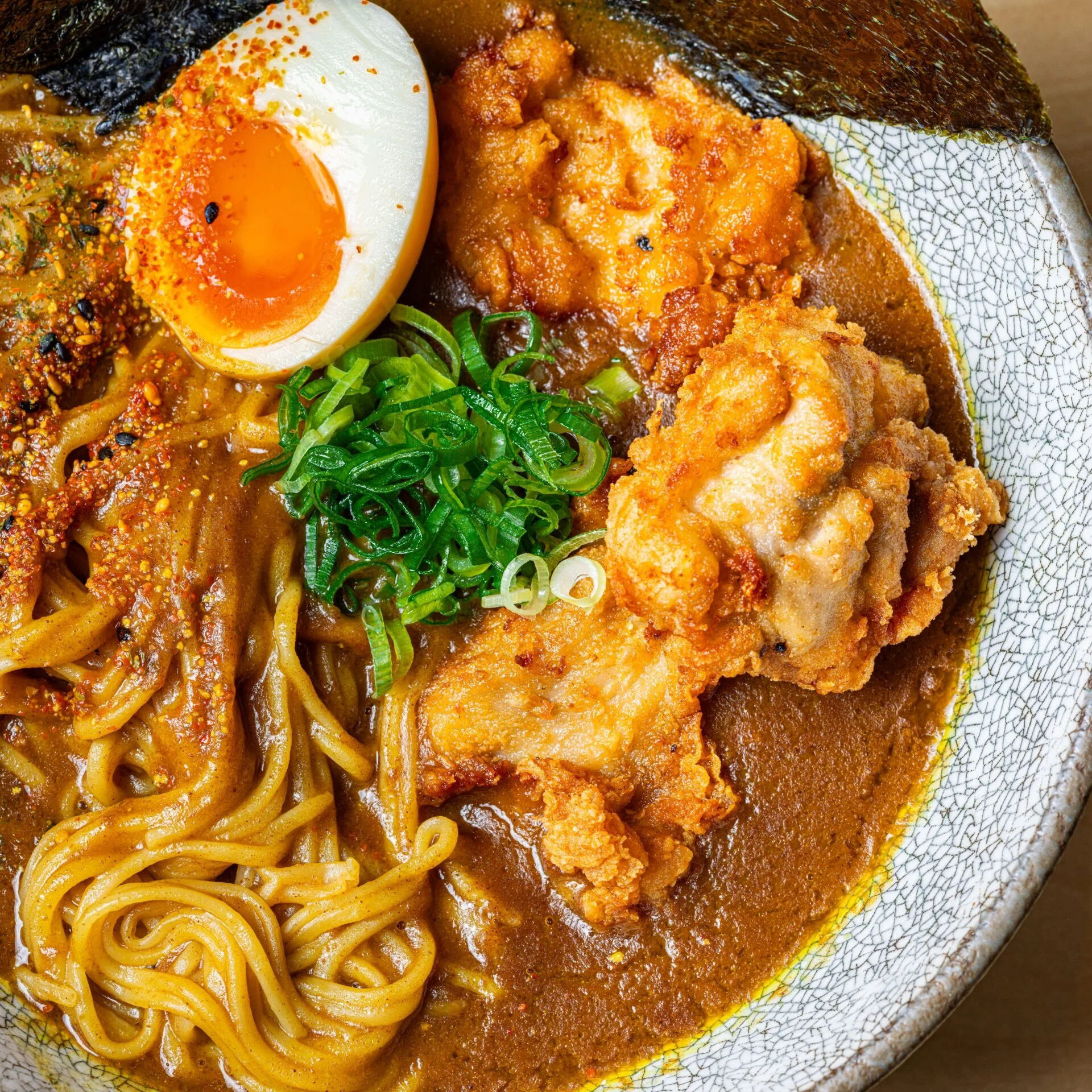 Close-up of a bowl of ramen with soft boiled egg, fried chicken, green onions, and spicy broth.