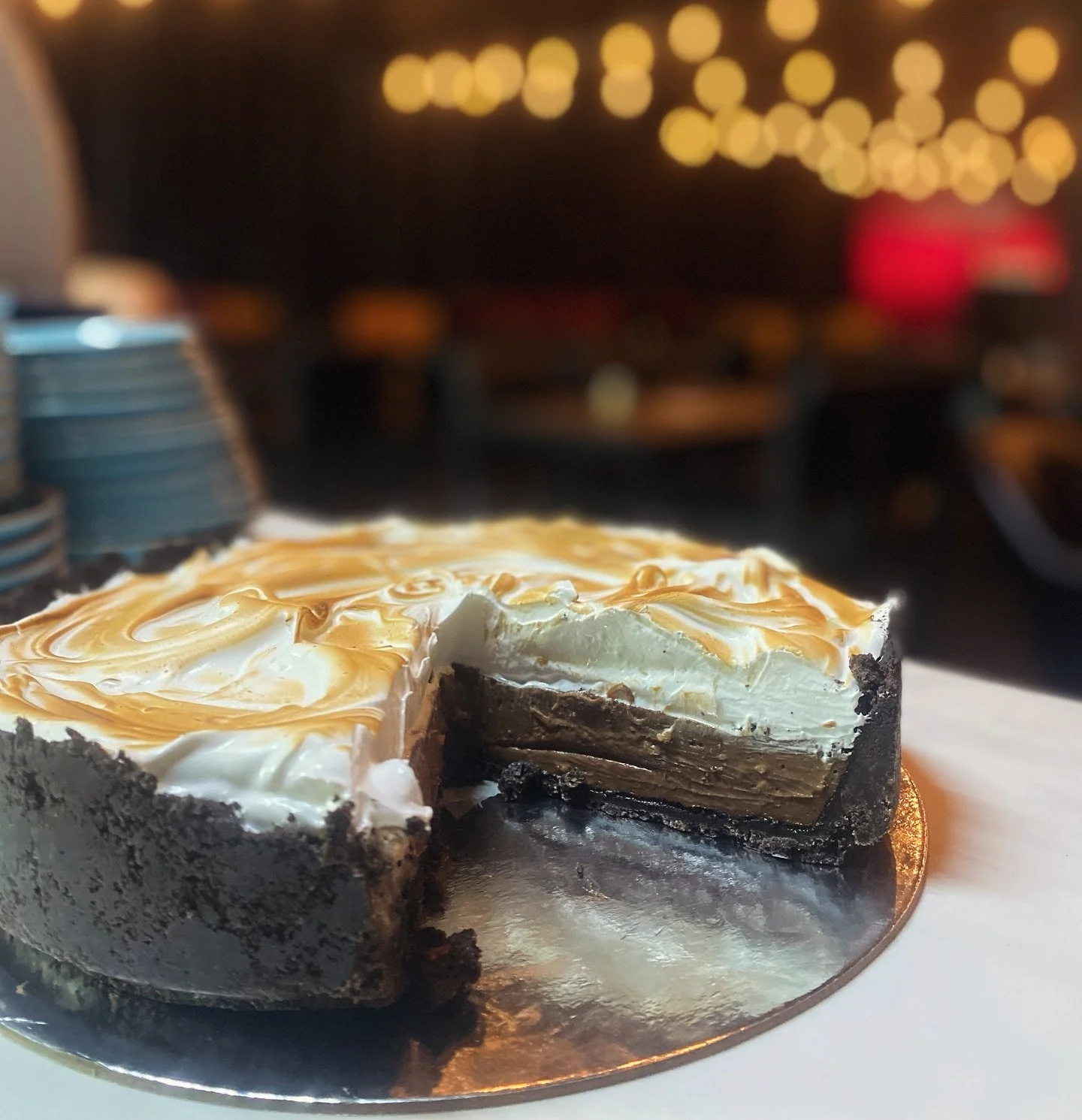 A pie with a chocolate crust, filled with cream and caramel, on a reflective silver surface with a slice removed, in a dimly lit setting.