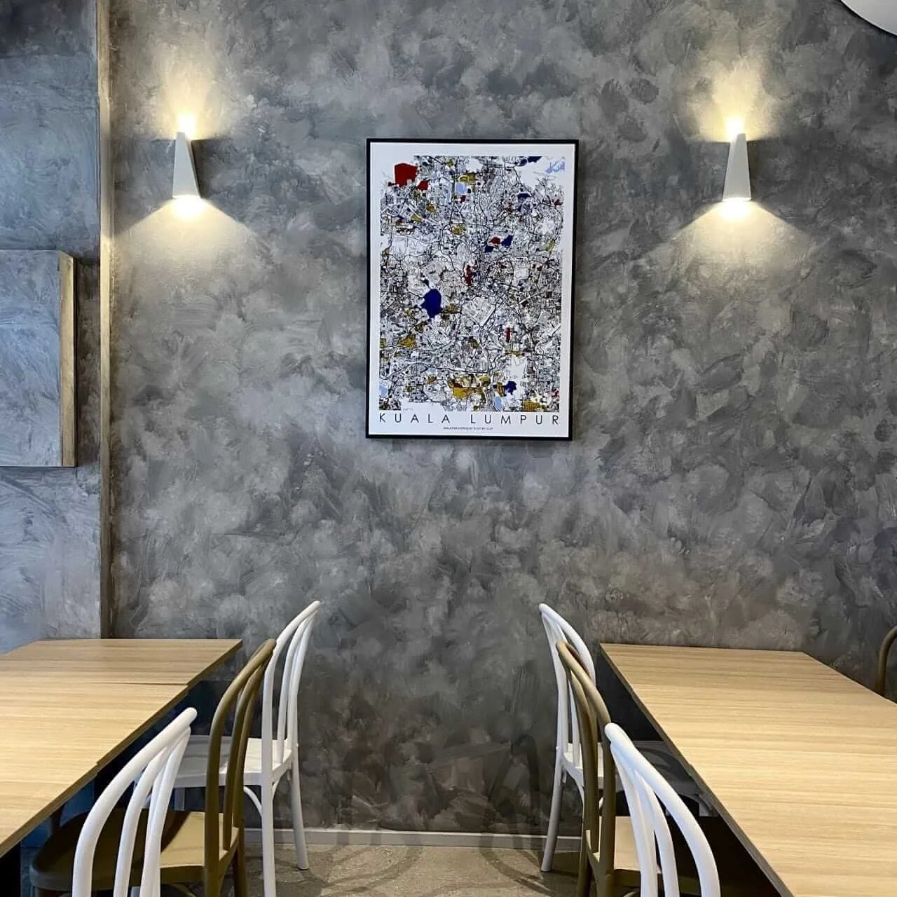 Interior of a restaurant with two tables, white and brown chairs, gray textured wall, and a framed map of Kuala Lumpur with wall sconces on each side.