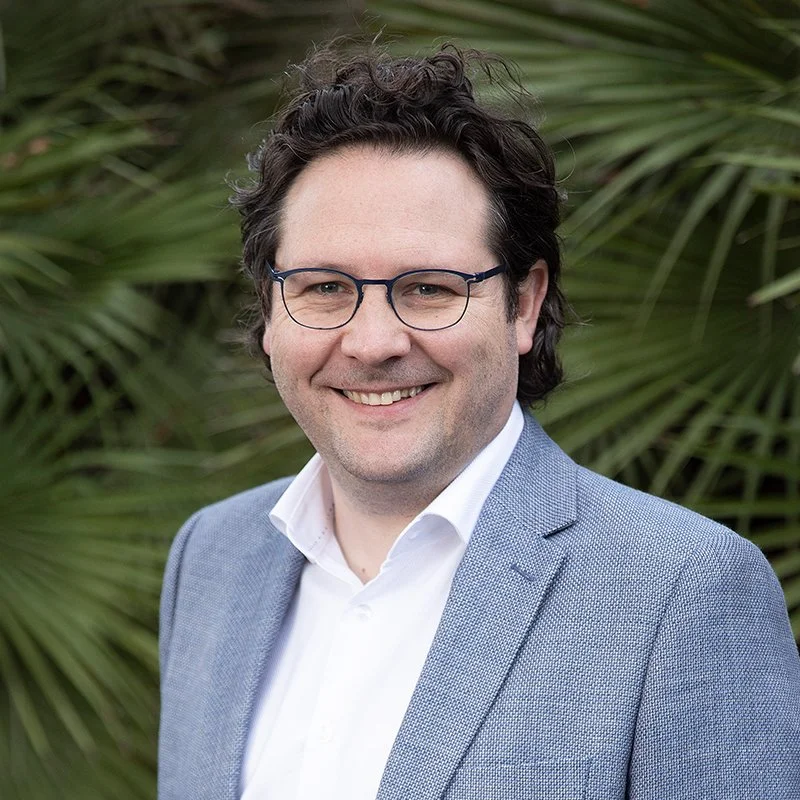 A man with curly dark hair, glasses, wearing a white shirt and light blue blazer, smiling outdoors with green palm leaves in the background.