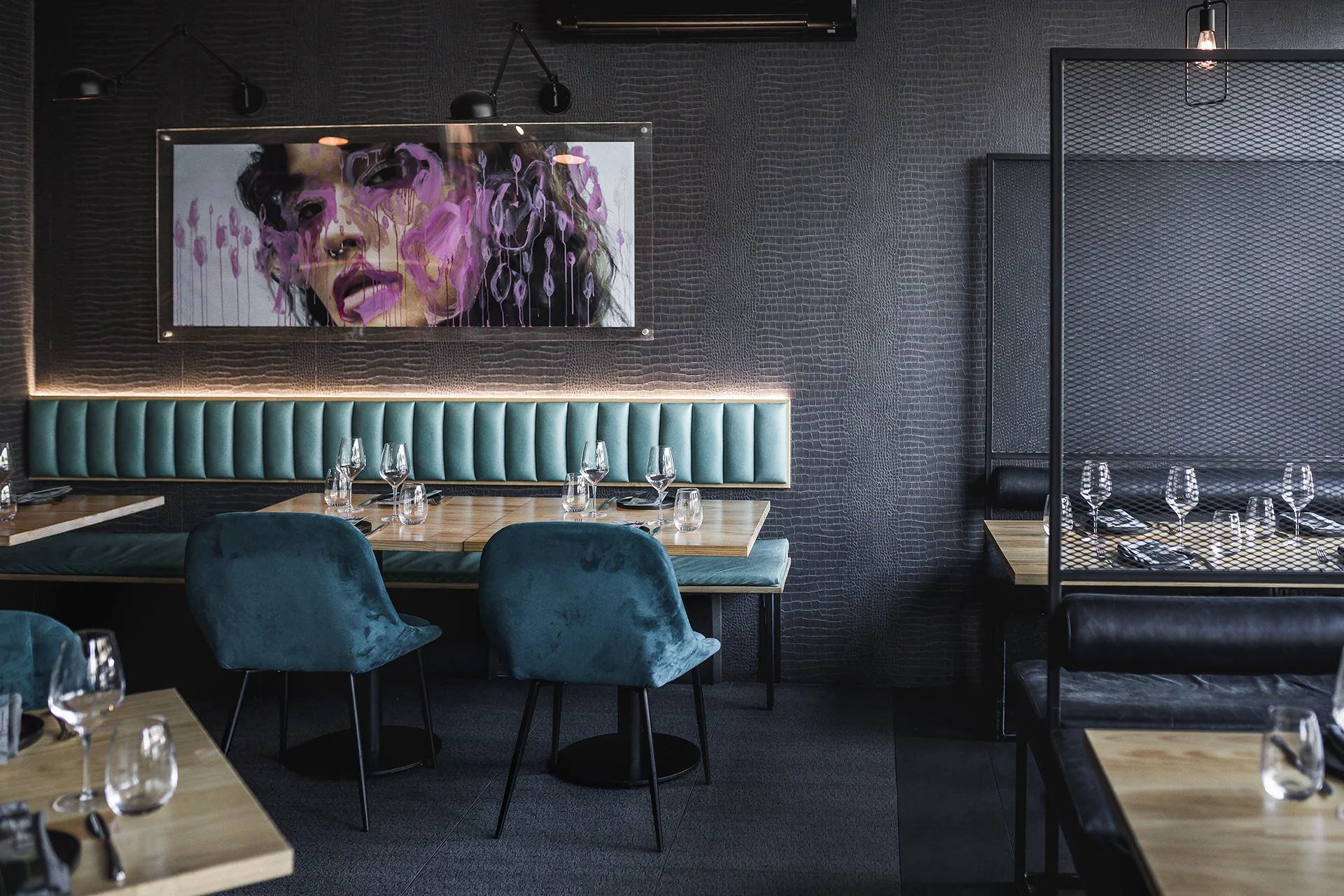 Interior of a modern restaurant with teal chairs and a padded teal banquette, decorated with glassware and table settings, dark textured walls, contemporary artwork of a woman's face with purple paint, and black lighting fixtures.