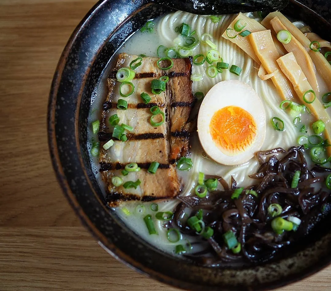 A bowl of ramen topped with grilled pork, half a soft-boiled egg, chopped green onions, bamboo shoots, and black fungus, on a wooden table.