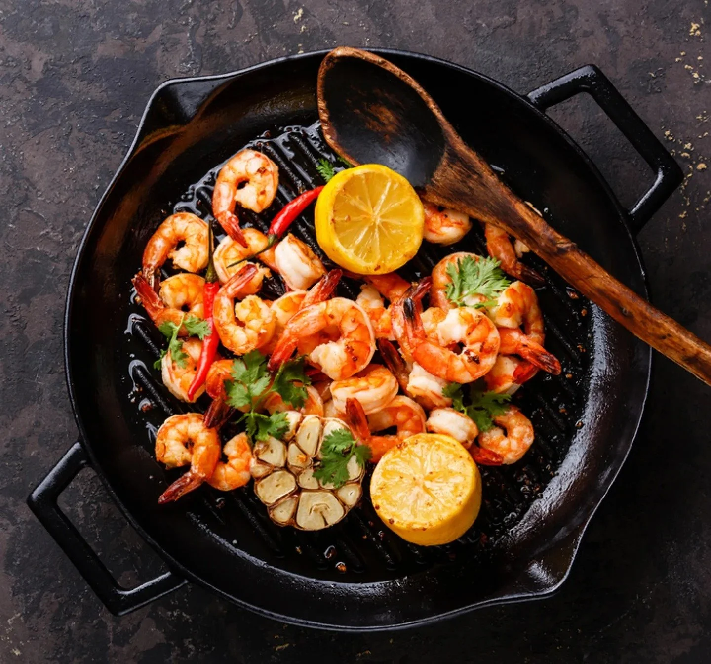 Shrimp and garlic being cooked in a black grill pan with lemon halves, red chili peppers, garlic, and cilantro garnishes.