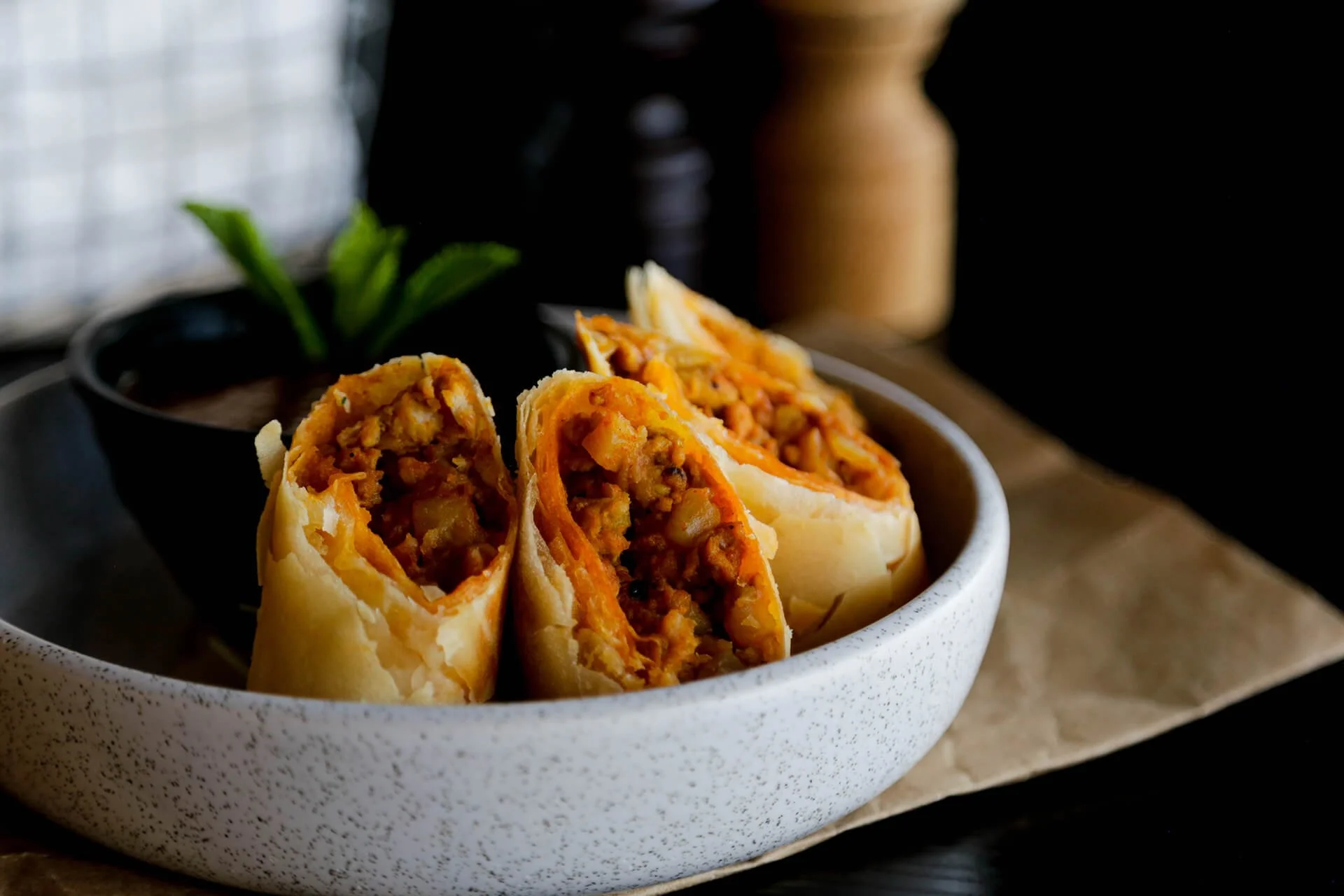 Close-up of a bowl of breakfast burritos filled with scrambled eggs, sausage, cheese, and potatoes, with a dark background and some green garnish in the background.