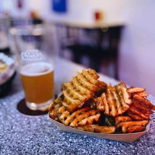 Basket of waffle fries with a glass of beer in the background on a speckled counter.