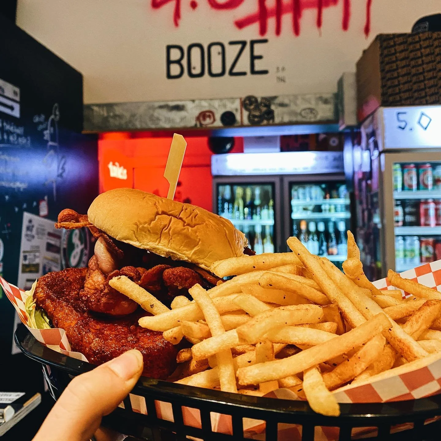 Basket of French fries with a fried chicken sandwich topped with crispy bacon, lettuce, and a sandwich bun, held by a person's hand in a restaurant or fast food setting.
