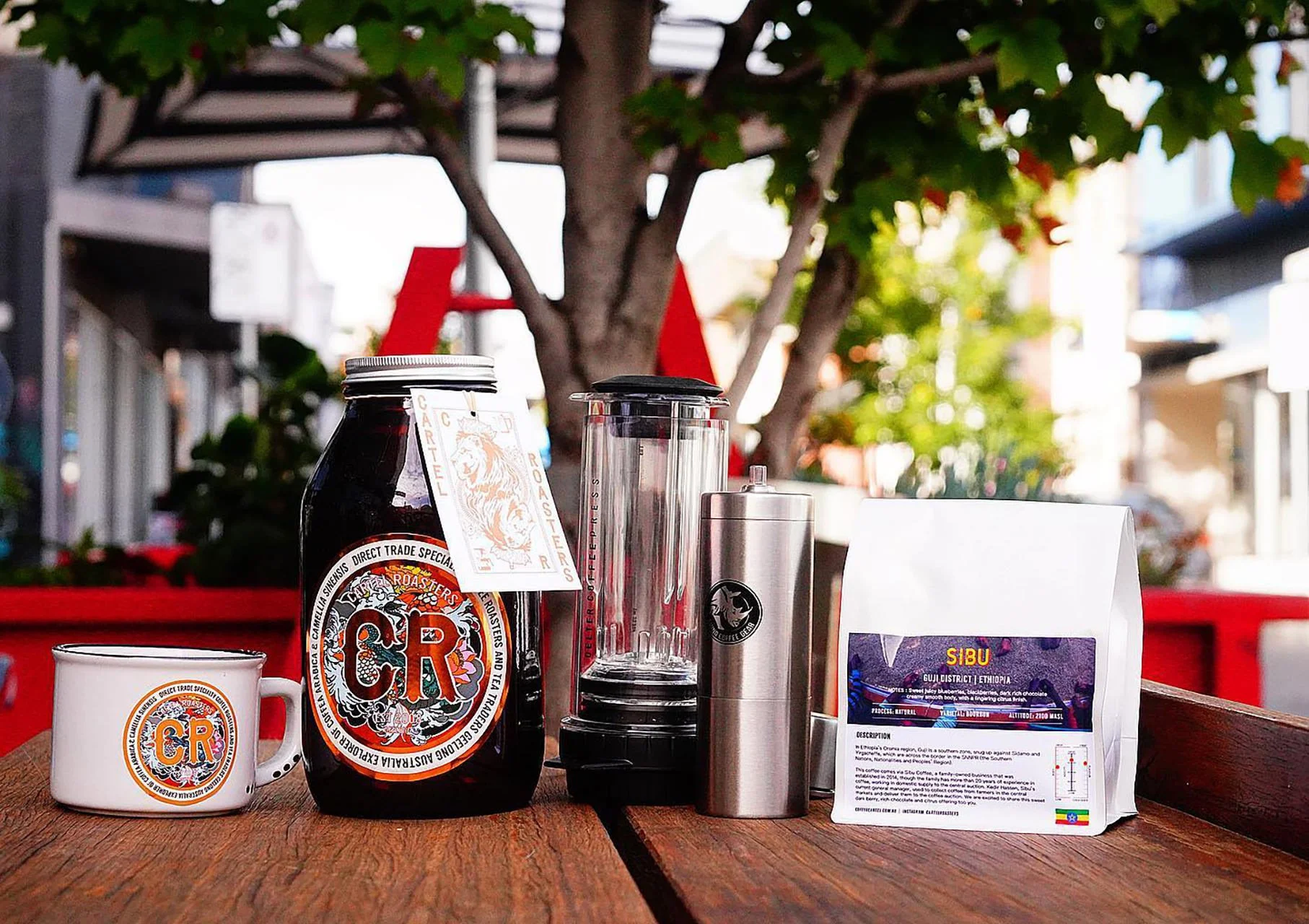 A coffee table with a ceramic mug, a large jar of tea labeled 'CR', a blender, a stainless steel travel mug, and a bag of Ethiopian Sibu coffee, outdoors with trees and buildings in the background.