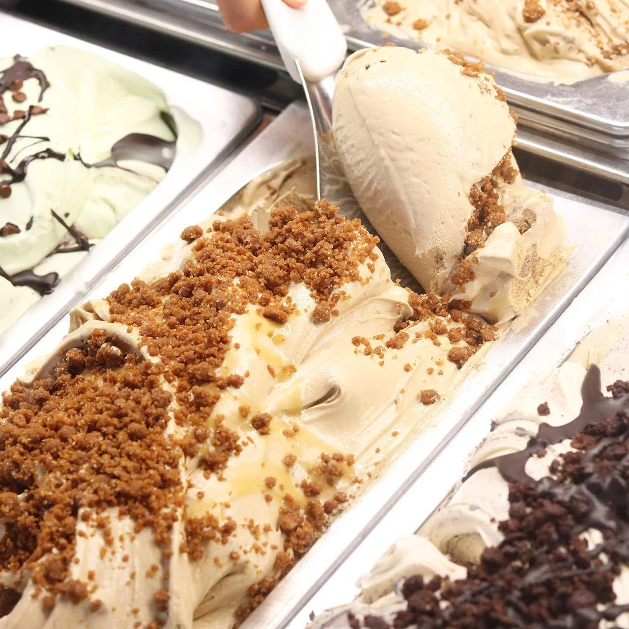 Ice cream in a metal tray being scooped into a cone, with caramel and crumb toppings, at a frozen dessert shop.