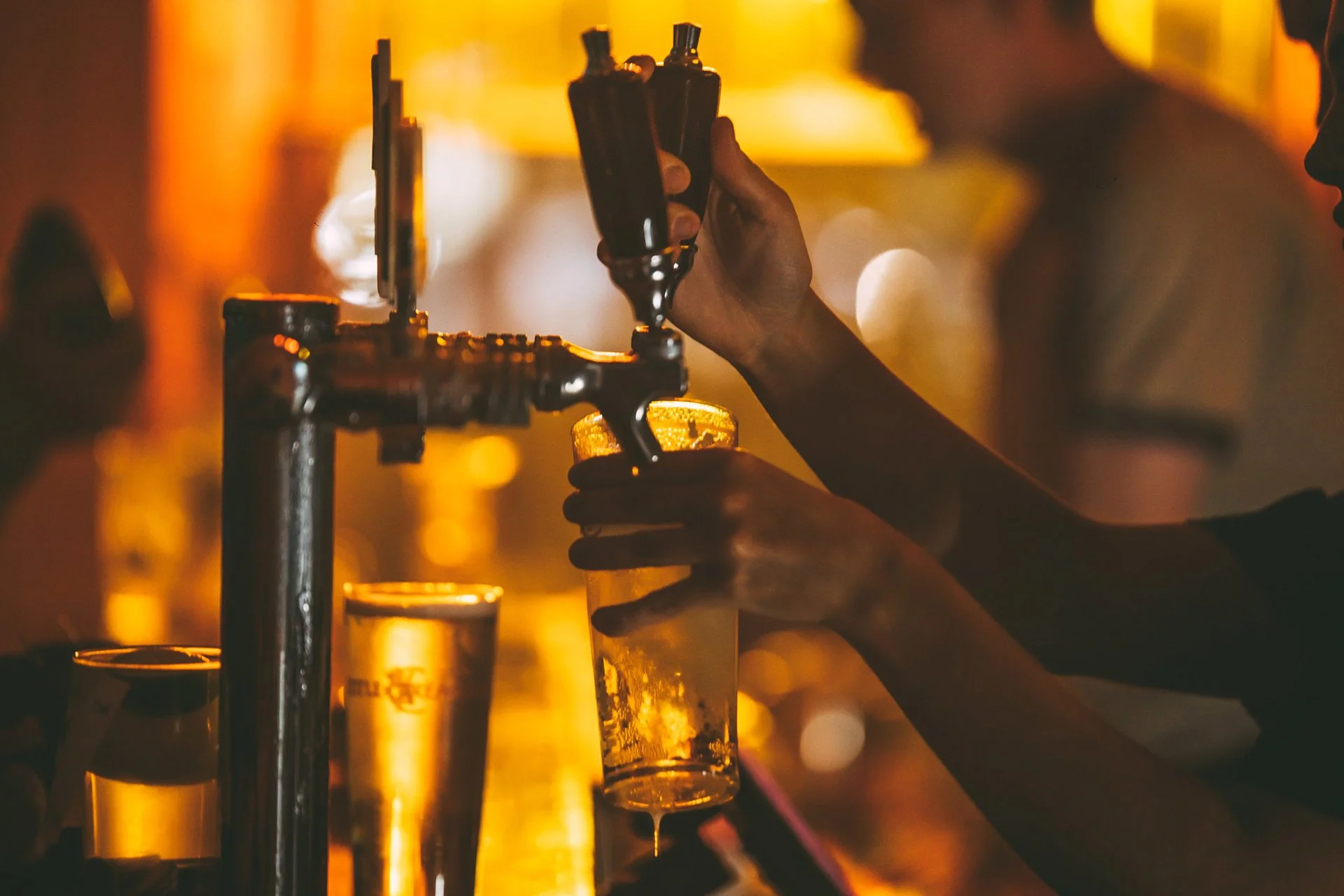 A bartender filming a beer glass below a beer tap in a dimly lit bar.